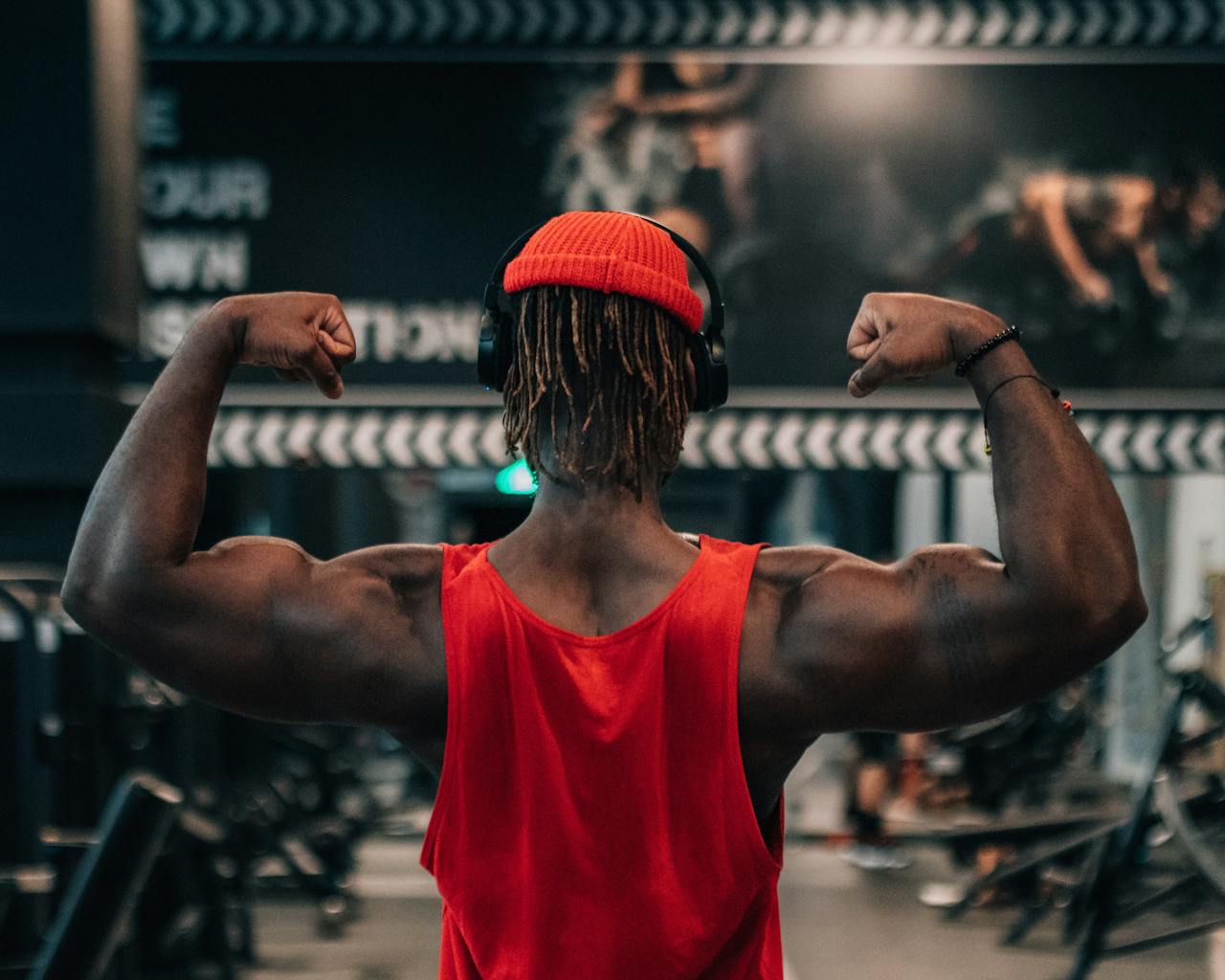 A man in a red tank top flexing his arm muscles in a gym setting, conveying motivation and strength.